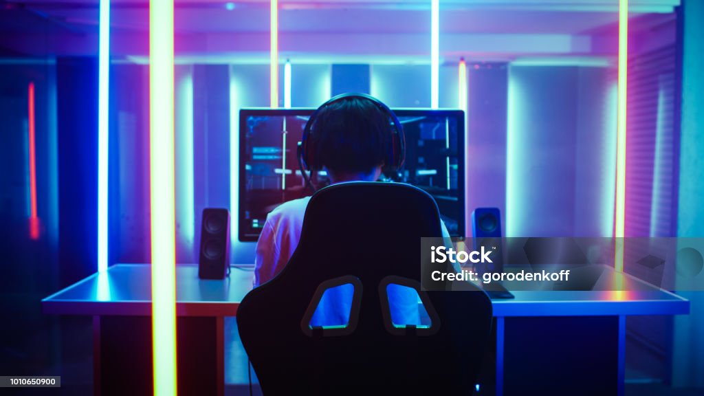 Back View Shot of the Beautiful Professional Gamer Girl Putting on Headset and Starts Playing Online Video Game on Her Personal Computer. Cute Casual Geek Girl. Room Lit by Neon Lamps in Retro Arcade Style. Video Game Stock Photo