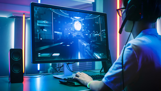 Back View Shot of Professional Gamer Playing in First-Person Shooter Online Video Game on His Personal Computer. He's Talking with His Team Through Headset. Room Lit by Neon Lights in Retro Arcade Style. Cyber Sport Championship.