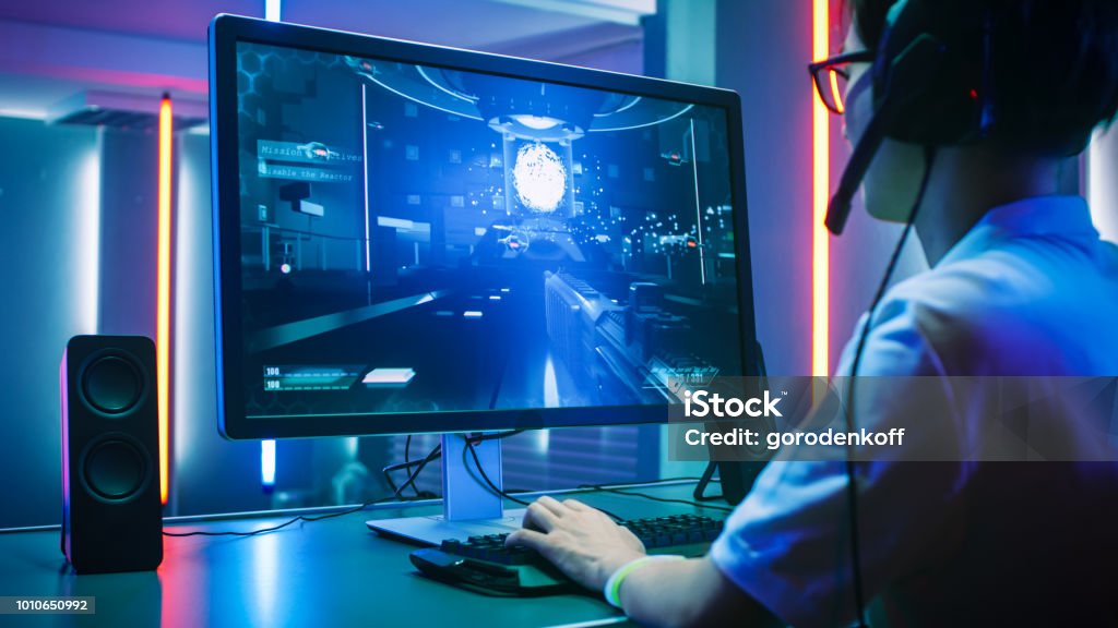 Back View Shot of Professional Gamer Playing in First-Person Shooter Online Video Game on His Personal Computer. He's Talking with His Team Through Headset. Room Lit by Neon Lights in Retro Arcade Style. Cyber Sport Championship. Video Game Stock Photo