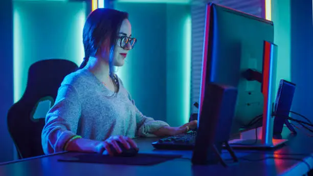 Beautiful Professional Gamer Girl Sitting Down to Play in First-Person Shooter Online Video Game on Her Personal Computer. Casual Cute Geek wearing Glasses and Talking into Headset. Cyber e-Sport Internet Championship.