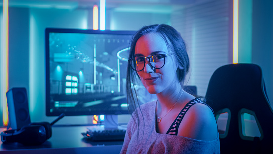 Retrato de la hermosa joven Pro Gamer, sentado frente a su computadora Personal y se ve en cámara. Jugador de chica atractiva Geek con gafas en la sala iluminada por luces de neón. photo