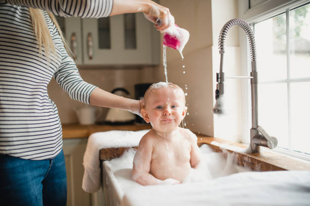Mother Pours Water Over Babies Head A small baby pulls a silly face in the sink as his mother pours warm water over his head to wash his hair. kitchen sink photos stock pictures, royalty-free photos & images