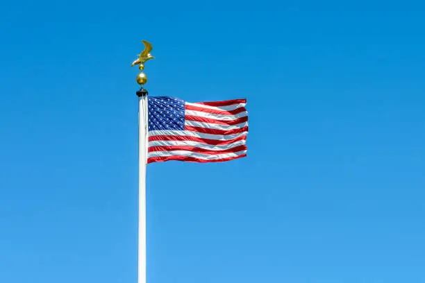Photo of The flag of the United States of America blowing in the wind at full-mast on a white pole topped with a golden eagle on ball ornament against deep blue sky.
