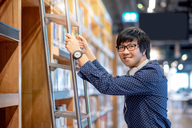 estudante jovem asiático escolhendo o livro da estante, usando a escada na biblioteca pública, pesquisador masculino com fones de ouvido e óculos fazendo pesquisa em universidade faculdade de educação e aprendizagem de conceitos - male education college student nerd - fotografias e filmes do acervo