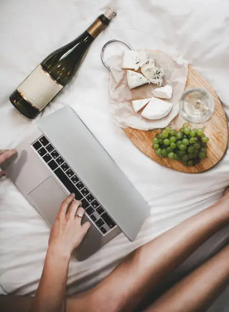 Grapes and bottle of white wine with cheese plate for the lunch in bedroom. Young woman is working at home