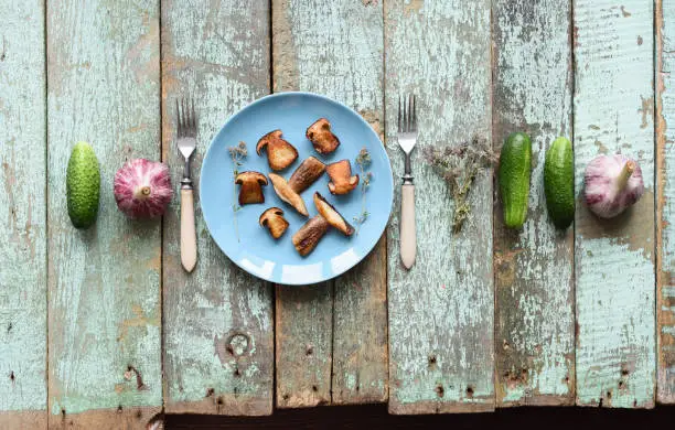 Nordic cuisine. Healthy vegan meal. Fried porcini (Boletus edulis) mushrooms in row with raw cucumbers, garlic and thyme on shabby blue background copyspace above view