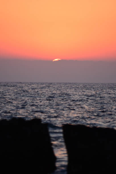 Sun setting behind distant clouds over the sea. In the foreground, a split boulder has separated giving a view of the sea . Photo taken in the Agios Georgios area of Cyprus. Nikon D7200 with Nikon 200mm macro lens