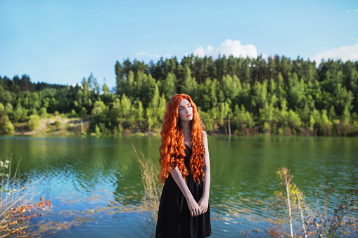 Young edwardian woman with long hair, red lips, pale skin on blue water background. Beautiful redhead model against the lake in black edwardian dress.