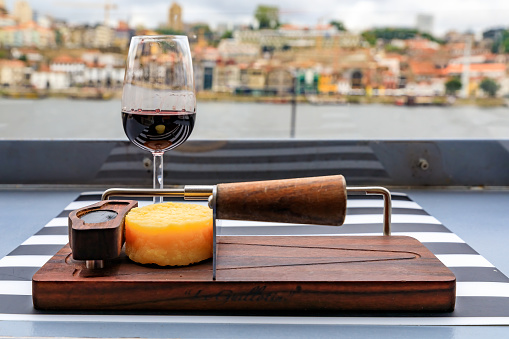 Glasses of port wine and a small wheel of cheese on the board at an outdoor restaurant with the Douro river and Dom Luis I bridge blurred in the background in Porto, Portugal