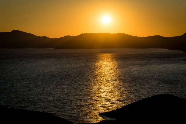 Setting sun, just before it dips below the mountains on the horizon, casts a beam of light across a bay of water at Torre di Pixinni, near Chia, Sardinia.