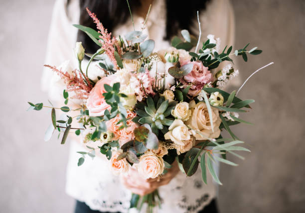 ótima jovem morena segurando o bouquet de flores noivas florescimento de rosas frescas, ranúnculo, suculentas, eucalipto em tons creme e rosa pastel sobre o fundo de parede cinza - wedding bride buying caucasian - fotografias e filmes do acervo