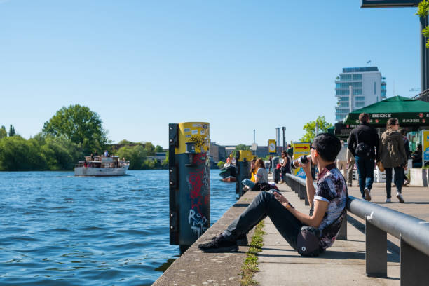 Berlin, Germany - may, 2018:  Young man drinks beer at riverside at river Spree, Berlin Wall (East side Gallery ) on a sunny day in Berlin, Germany