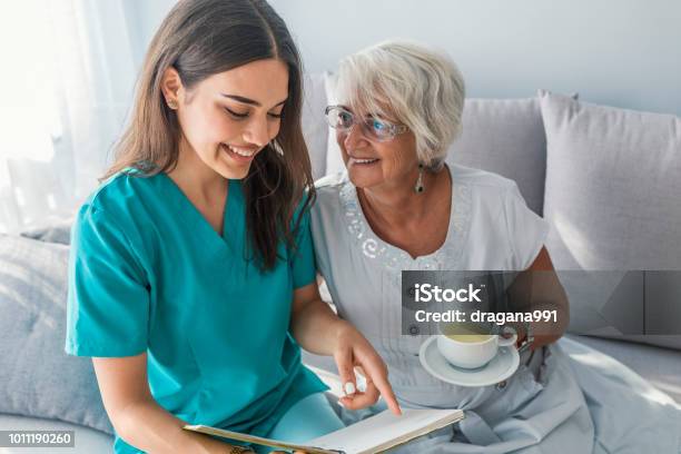 Happy Elder Woman Sitting On White Sofa And Listening To Nurse Reading A Book Out Loud Stock Photo - Download Image Now
