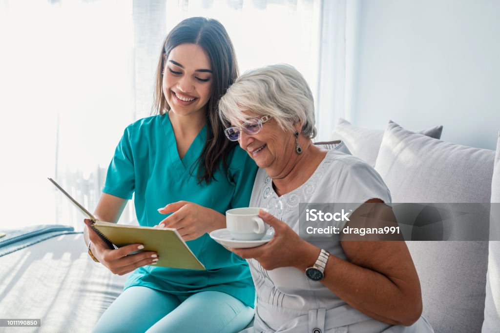 Donna che legge un libro mentre è seduta con la nonna felice che beve il tè - Foto stock royalty-free di Infermiera - Infermiere