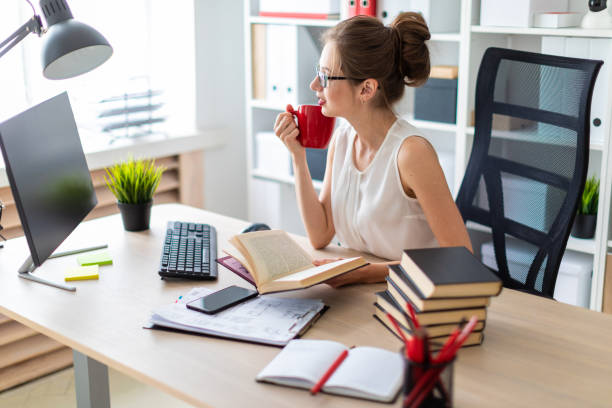 istock A young girl sits at a computer table and holds an open book and a red cup in her hands. 1011251006