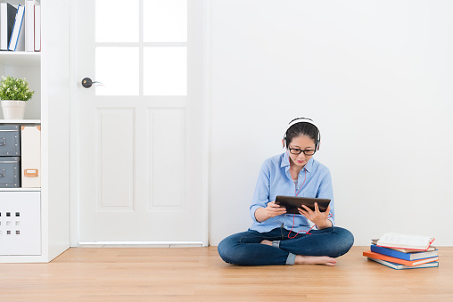 young university female student sitting on wood floor using mobile digital tablet looking at online video relaxing when she finished doing school homework.