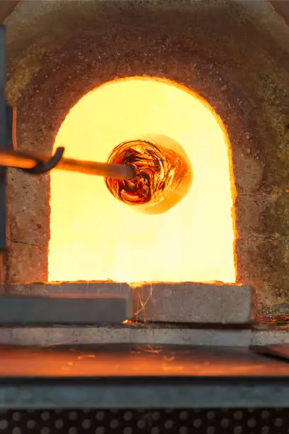 Photo of Murano glass-blowing factory. Glass blower forming beautiful piece of glass: put iron rod with attached glass object in furnace to make the glass malleable. Italy