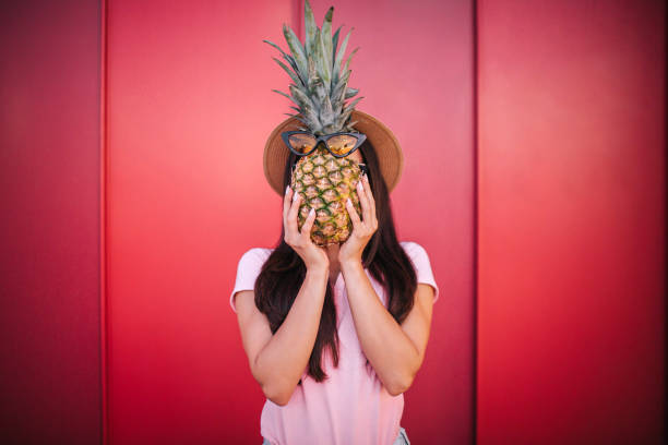 istock Girl stands and covers her face with pineapple. She holds it with both hands. Isolated on striped and red background 1011263210