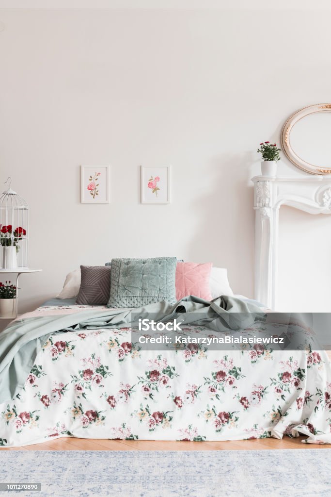 Two simple pink posters hanging on wall above double bed with cushions and floral sheets in real photo of white bedroom interior Bedroom Stock Photo