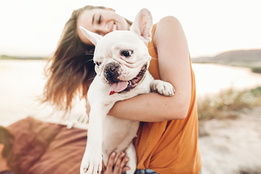 happy hipster woman playing with bulldog on the beach in sunset light, summer vacation. stylish girl with funny dog resting, hugging and having fun in sun, cute moments. space for text