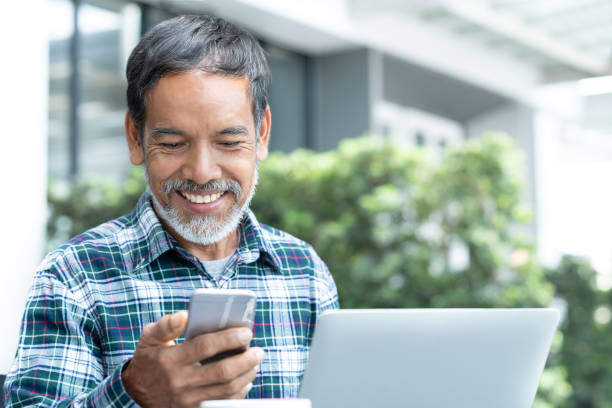 istock Smiling happy mature man with white stylish short beard using smartphone gadget serving internet at coffee shop cafe outdoor. Laughing old man using social media network technology feeling excited. 1011293604