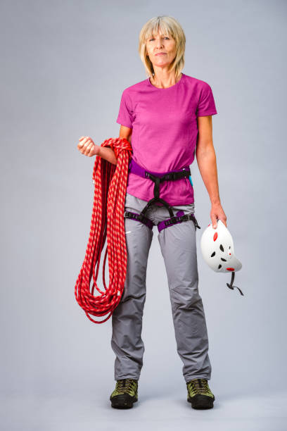 Senior woman standing with rock climbing equipment against a grey background.
