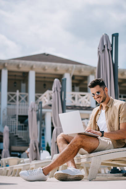 istock smiling young man in sunglasses sitting on chaise longue and using laptop 1011331804