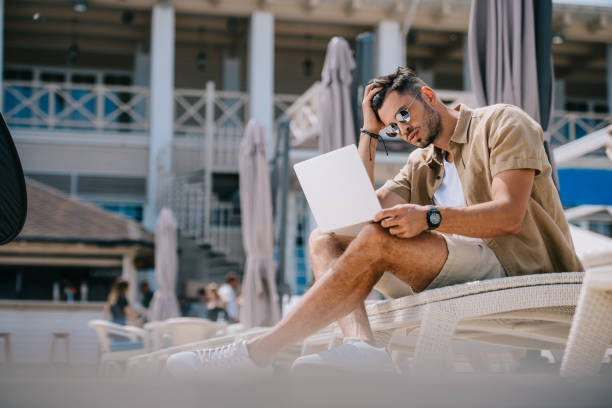 istock handsome young man in sunglasses using laptop while resting on chaise longue 1011337066