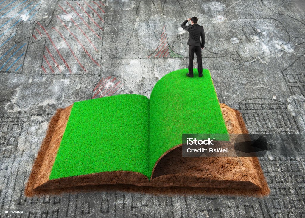 Businessman standing on open book of grass and soil texture Small businessman standing on the opened book of green grass and soil textured, on dirty doodles concrete floor background, concept of ECO, renewable energy and circular economy. Circular Economy Stock Photo