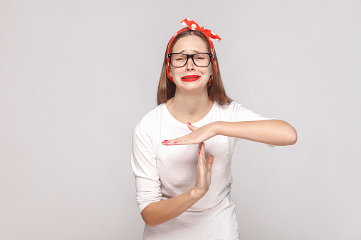 timeout, please give me more time. portrait of beautiful emotional young woman in white t-shirt with freckles, black glasses, red lips and head band. indoor shot, isolated on light gray background.