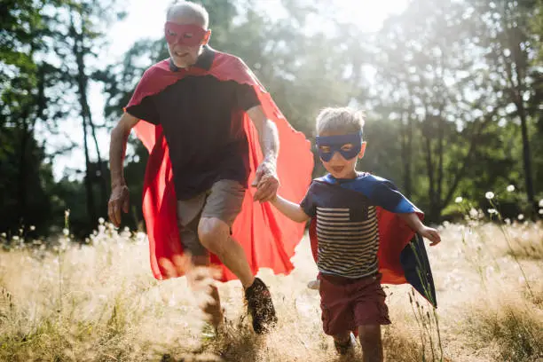 A senior man has fun dressing up in a hero mask and cape costume with his grandchild, a boy about 4 years old.  They have fun playing a running around in a sunny grassy field.