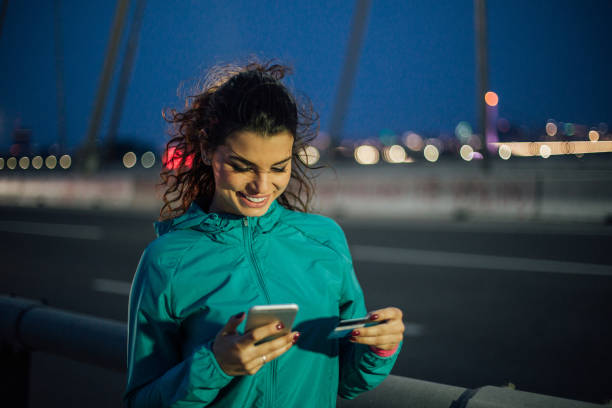 Fit young woman shopping online after outdoor night exercise Young woman using credit card and smart phone charging sports photos stock pictures, royalty-free photos & images