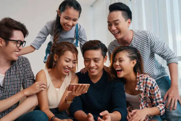 Group of joyful laughing young people watching funny videos on smartphone screen at home