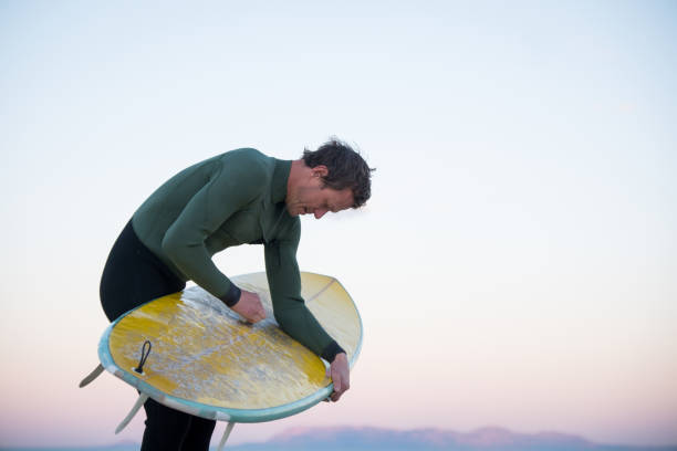 Man gets ready to go for an early morning surf, he is waxing his surf board.