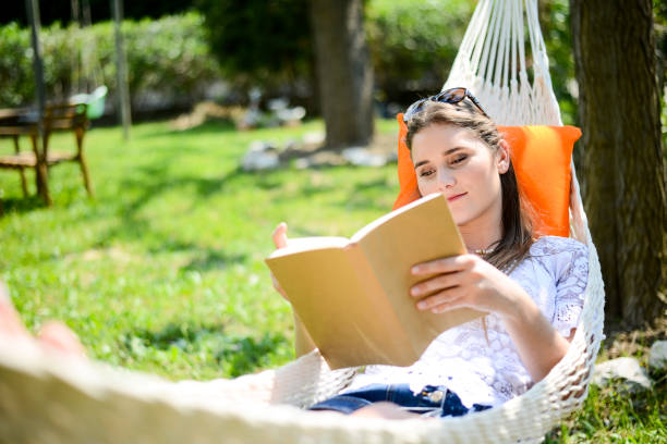 pretty young woman relaxing and reading a book in hammock outdoor in the garden during a sunny summer day pretty young woman relaxing and reading a book in hammock outdoor in the garden during a sunny summer day hammock relaxation women front or back yard stock pictures, royalty-free photos & images