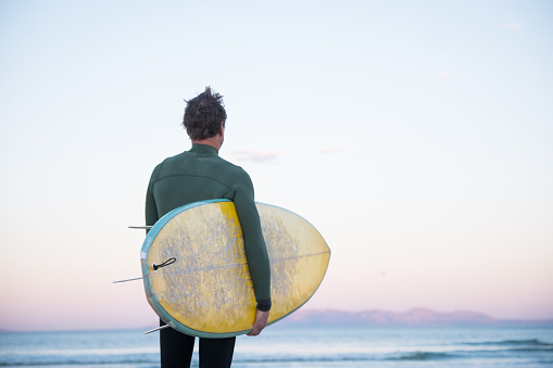 Young man walks down the beach with his surfboard in his hand ready to go and surf in False bay, the sun is rising over the horizon.