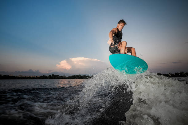 uomo attivo e forte che salta sul wakesurf lungo le onde del fiume al tramonto - wakeboarding surfing men vacations foto e immagini stock