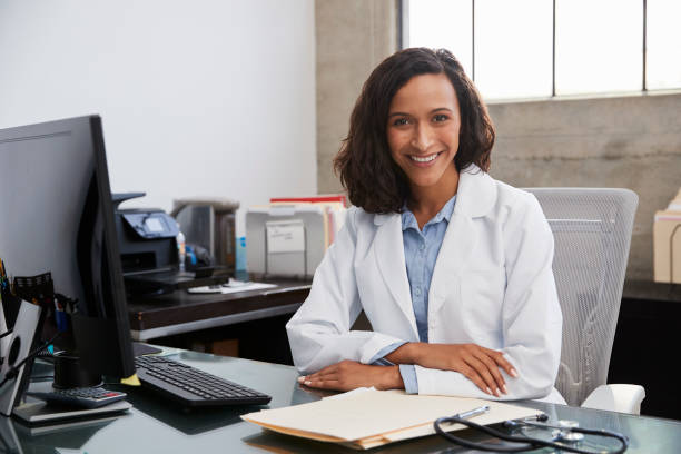 young female doctor sitting at desk in an office, portrait - therapy people cheerful looking at camera imagens e fotografias de stock