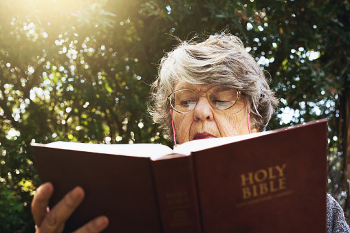 An old woman reads the Holy Bible in her leafy garden looking serious.