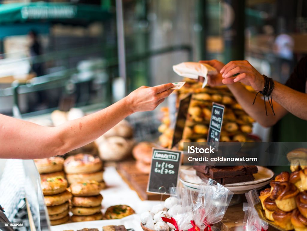 Customer paying for sweet pastry using cash at food market Close up color image depicting a cash transaction between a customer and the owner of a bakery stall at an outdoor food market in London, UK. The customer is paying using a five pound note for what appears to be some kind of cake or pastry. In the background the contents of the market stall - cakes, brownies, sweet pastries - are blurred out of focus. Room for copy space. Food Stock Photo