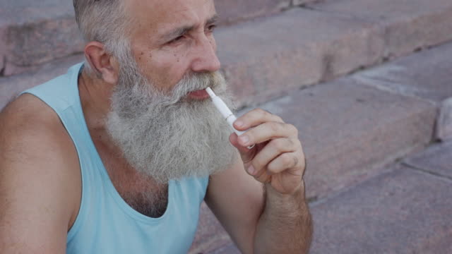 bearded senior man sitting on the steps and smokes an e-cigarette