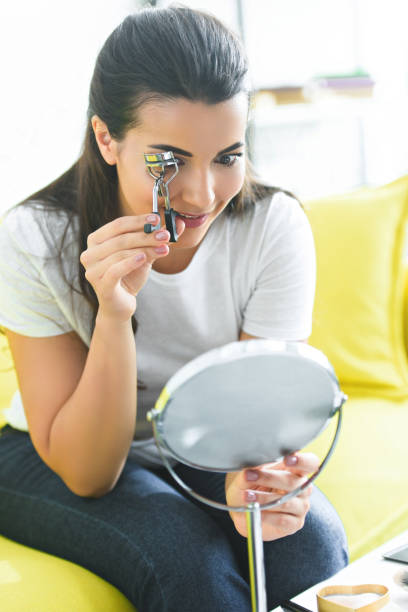 istock young woman looking at mirror while curling eyelashes with eyelash curler at home 1011964708