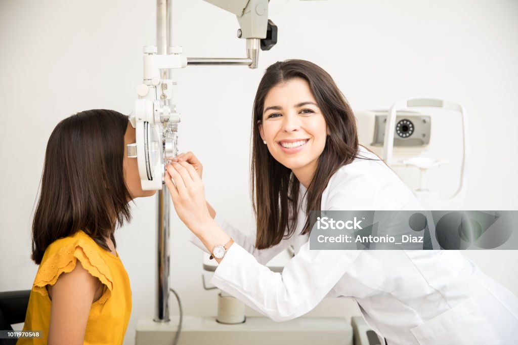 Confident optometrist examining patient eyes Portrait of happy beautiful woman doing eye test of girl child in optical store while looking at camera Ophthalmologist Stock Photo