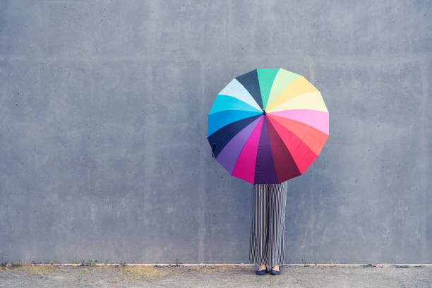 young woman standing with multicolored umbrella in front of a concrete wall - rainbow umbrella descriptive color multi colored imagens e fotografias de stock