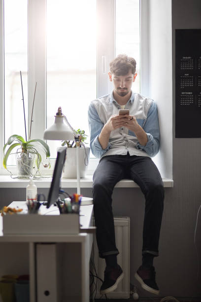 istock Young stylish man sitting on the window with phone in hands 1012020086