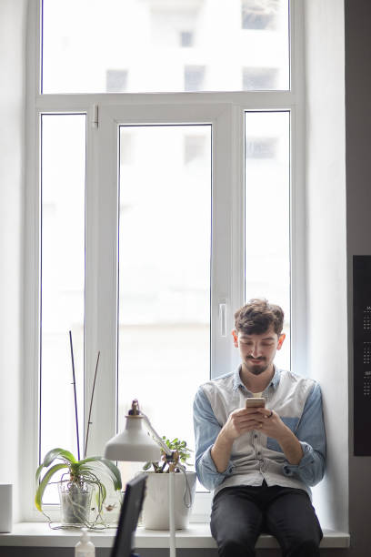 istock Young stylish man sitting on the window with phone in hands 1012020168