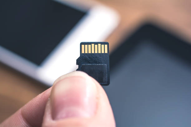 Male Fingers Holding A Micro SD Card With Tablet And Smartphone In Background, Extent Disk Space Concept stock photo