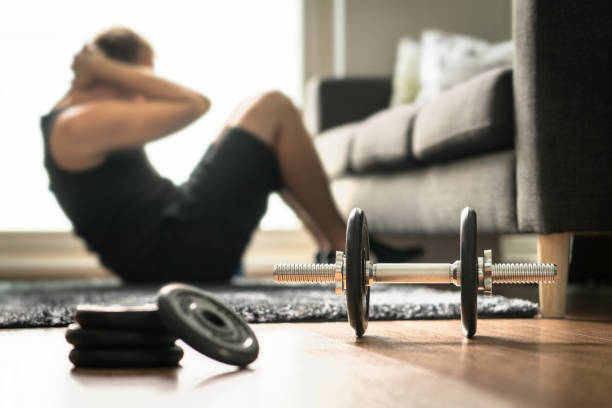 istock Home workout. Man doing ab training and crunches in living room gym. Guy doing sit ups. Warm up before weight exercise. Fitness concept with dumbbell. 1012049110