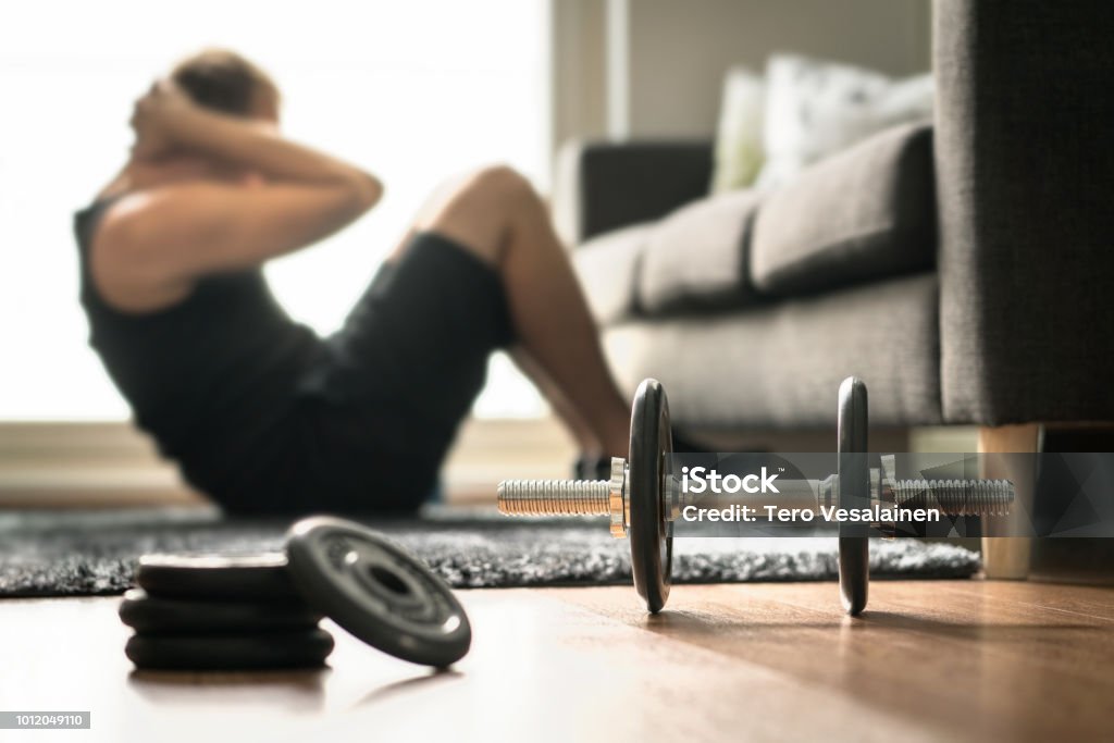 Home workout. Man doing ab training and crunches in living room gym. Guy doing sit ups. Warm up before weight exercise. Fitness concept with dumbbell. Home workout. Man doing ab training and crunches in living room gym. Guy doing sit ups. Warm up before weight exercise. Fitness concept with dumbbell and athlete. Exercising Stock Photo