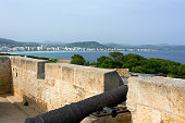 View of an antique iron cannon on the old defence tower of Punta de N'Amer near Sa Coma, with the seaside resort Cala Millor in the background on the Spanish Balearic Mediterranean island of Mallorca